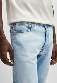 Jeans din denim de un albastru deschis au o croială slim și o ușoară decolorare. Acestea au un design cu cinci buzunare, cu cusături contrastante și un mic etichetă roșie.