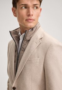 Beżowa marynarka z mieszanki wełny z ostrym klapem, na brązowej, pikowanej kurtce zapinanej na zamek. Charakterystyczne detale to kieszonka na piersi oraz fakturowana tkanina.