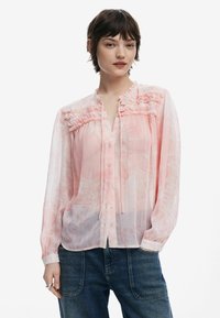 Blusa rosa chiaro con dettagli arricciati sulle spalle, chiusura con bottoni e maniche lunghe, abbinata a jeans in denim scuro.