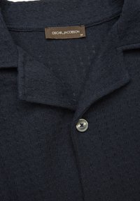 Granatowy sweter dzianinowy z kołnierzykiem, srebrnym guzikiem i teksturowaną tkaniną z subtelnym wzorem. Widoczna metka marki.