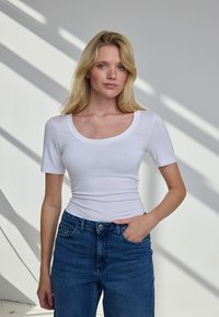 Blondynka w białym dopasowanym krótkim rękawie i niebieskich dżinsach, stojąca z jedną ręką w kieszeni na tle białej ściany z cieniami.