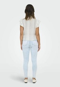 Haut blanc en dentelle avec un dos en dentelle et des manches à volants, associé à un jean skinny bleu clair et des chaussures claires. Motif de dentelle détaillé au dos.