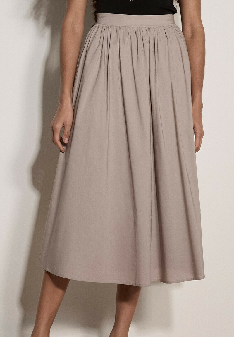Jupe midi beige avec une taille haute et des plis, portée par une personne debout devant un fond clair uni.