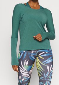 Långärmad, grön atletisk topp med en avslappnad passform. Kombinerad med leggings som har ett tropiskt växtmönster i blått, gult och vitt.