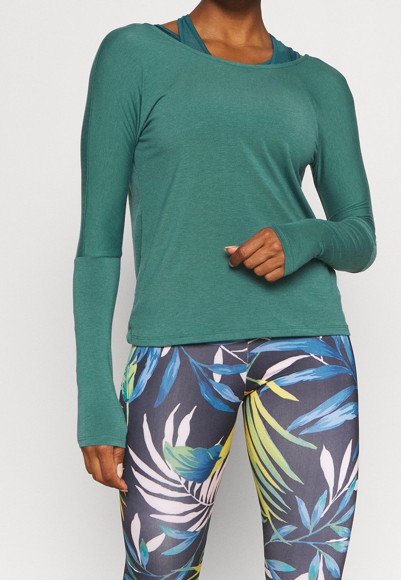 Långärmad, grön atletisk topp med en avslappnad passform. Kombinerad med leggings som har ett tropiskt växtmönster i blått, gult och vitt.