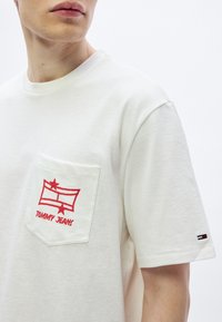 Camiseta blanca de algodón con bolsillo que presenta un diseño gráfico rojo y texto "TOMMY JEANS". Mangas cortas y un pequeño logo en la manga.