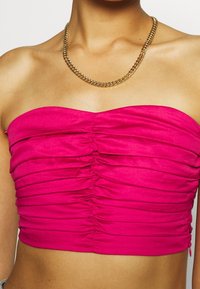 Kobieta w jasnoróżowej, marszczonej bluzce bez ramiączek oraz złotej naszyjniku, odsłaniająca ramiona i górną część klatki piersiowej.