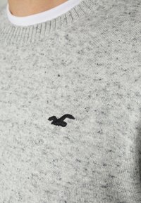 Šedý pletený svetr s tečkovanou texturou. Má vroubkovaný kulatý výstřih a malý černý logo na hrudi. Měkký materiál.