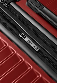 Plano detalle de la cremallera de una maleta rígida roja con ribete negro y una etiqueta de tirador que dice "REDOLZ".