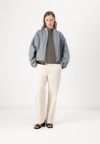 Giacca bomber blu chiaro con una cerniera argentata, indossata sopra un dolcevita grigio scuro e pantaloni a gamba larga color crema, accentuata con sandali neri con tacco.