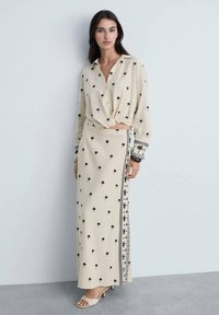 Blouse beige avec motifs de palmiers noirs, nouée à la taille ; jupe longue assortie avec des motifs similaires et une garniture décorative sur le côté. Sandales plates.
