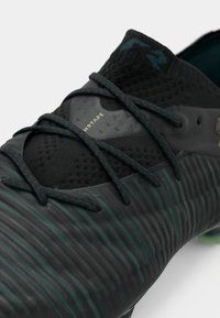 Close-up of black and dark green textured soccer cleat with laces and the word "WRTAE" on the upper fabric section.