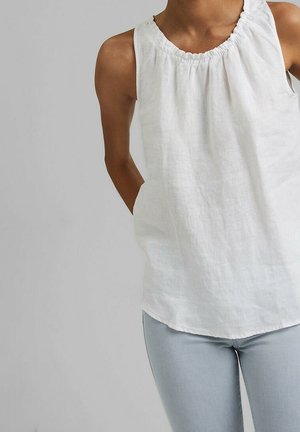 Frau trägt ärmellose weiße Leinenbluse mit gerafftem Ausschnitt und hellblaue Jeans, steht vor einfachem grauem Hintergrund.