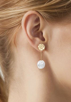 Close-up van een oor met een goud-witte hangende oorbel tegen een achtergrond van een lichte huid en blond haar.