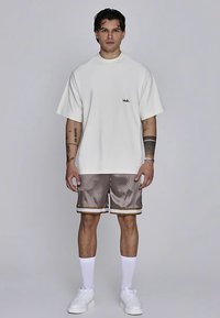 Valge üle suurusega t-särk, rinnal logo, kombineeritud pruunide satiinist lühikeste pükstega, kuldsed detailid, valged sokid ja valged tossud. Lihtne taust.