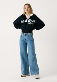 Skraćeni crni dukserica s logom "Red Bull Racing", u kombinaciji s širokim svjetloplavim trapericama i bijelim tenisicama sa sivim detaljima.