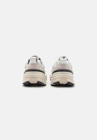 Un paio di sneakers beige e bianche viste da dietro, che mostrano suole spesse e pannelli del tallone testurizzati su uno sfondo semplice.
