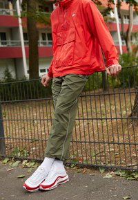 Röd nylonjacka med framficka, olivgröna smalna byxor med vita detaljer och vita/röda sneakers med luftkuddar. Avslappnad utomhusmiljö.