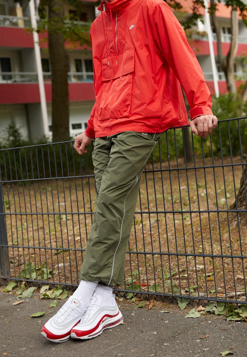 Röd nylonjacka med framficka, olivgröna smalna byxor med vita detaljer och vita/röda sneakers med luftkuddar. Avslappnad utomhusmiljö.