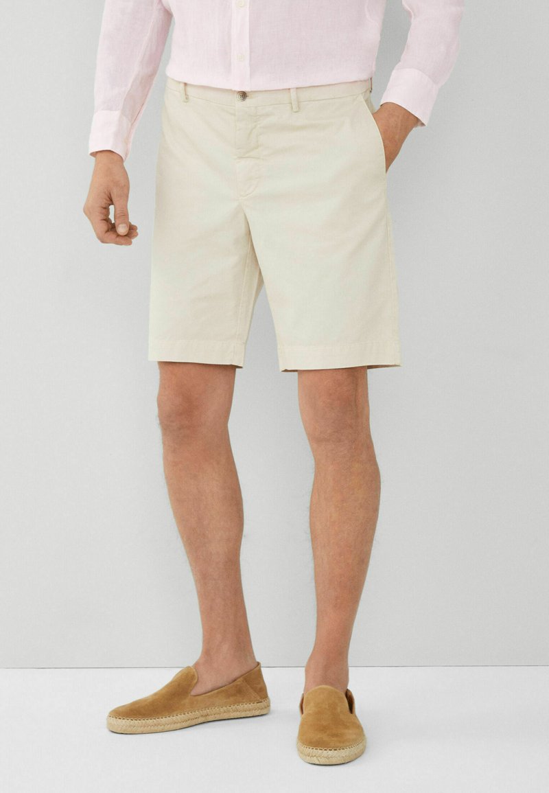 Beige bomulds shorts med en skræddersyet pasform, med flad front, sidelommer og synlige sting. Kombineret med brune slip-on sko.