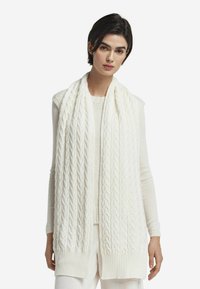 Hvid strikket tørklæde med kabelmønster og ribkanter, elegant draperet over en lyse farvet top, der fremhæver dets teksturerede design.