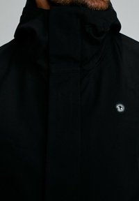 Homme portant une veste noire à col montant zippée, avec un petit logo circulaire sur la poitrine.