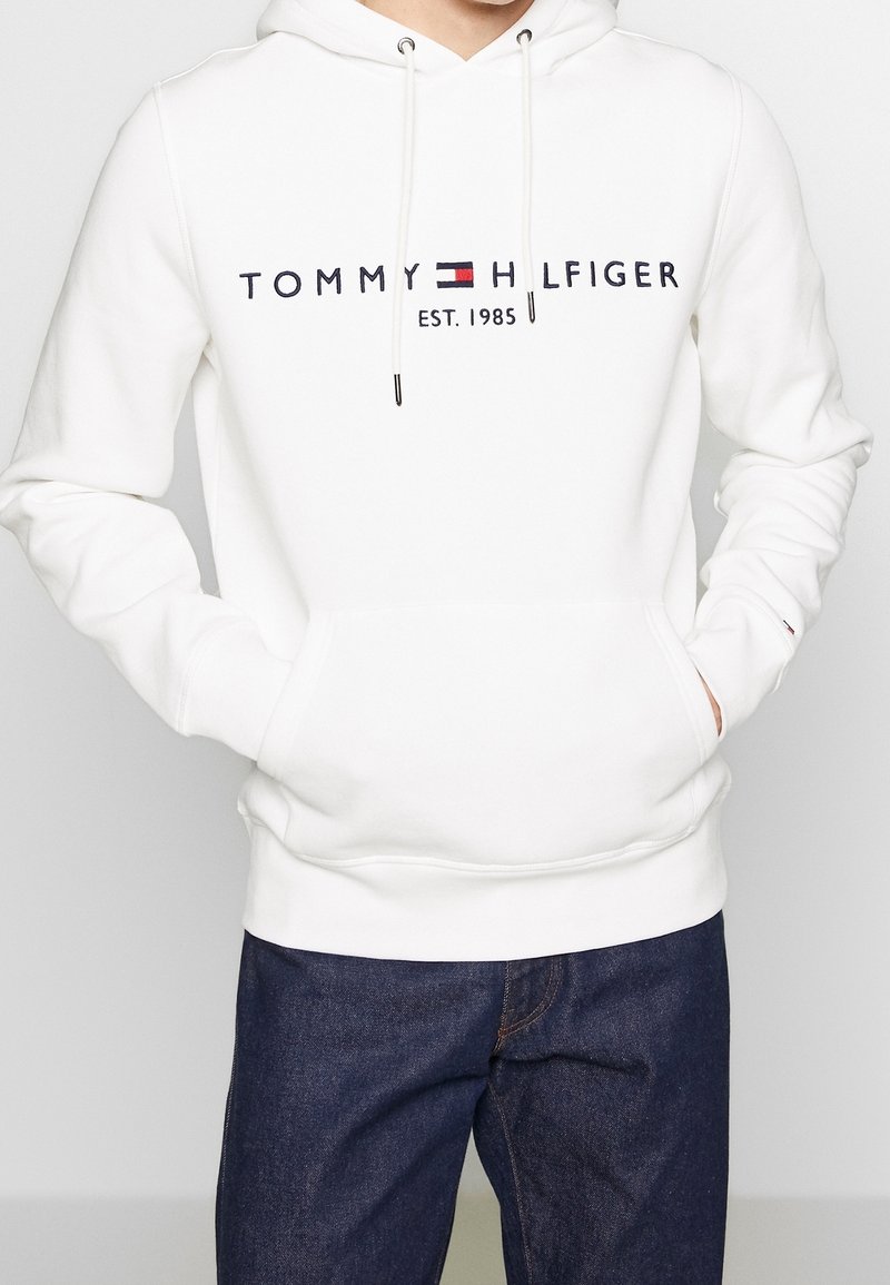 Felpa bianca con cappuccio e coulisse regolabili, con la scritta "TOMMY HILFIGER" e "EST. 1985" stampati in blu scuro, polsini a costine e una tasca frontale.