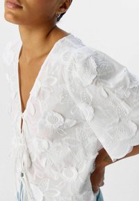 Hvid broderet bluse med blomsterpræg, en dyb V-hals og pufærmer. Stoffet har en let, gennemsigtig tekstur med bindebånd.