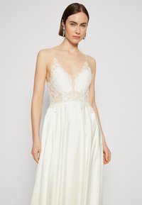 Vit klänning med djup V-ringning och genomskinlig spetskorsett med blommönster. Kjolen är slät och flödar elegant, vilket betonar silhuetten.