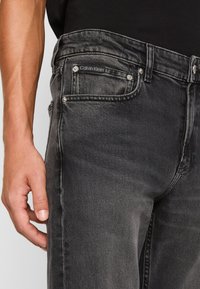 Jeans de mezclilla negra con una textura descolorida, que presentan una etiqueta de Calvin Klein en el bolsillo delantero y detalles de herrajes metálicos. Corte ligeramente entallado.