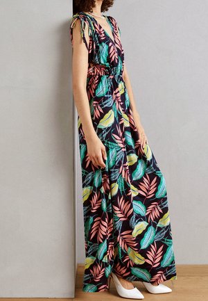 Femme portant une robe maxi noire sans manches avec un imprimé coloré de feuilles tropicales et des talons blancs pointus, appuyée contre un mur.