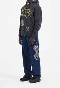 Felpa grigia con cappuccio con grafica di teschio e testo; jeans blu in denim con ricamo colorato di drago; scarpe da ginnastica nere.