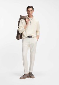 Άνδρας που φοράει κρεμ παντελόνι, ανοιχτό μπεζ πουλόβερ με φερμουάρ, ταμπά αθλητικά παπούτσια και κρατάει μια ταμπά ζακέτα στον ώμο του, στέκεται σε λευκό φόντο.