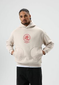 Beige hoodie met een ronde rode afbeelding met een roosontwerp en tekst. Ronde, oversized zakken aan de voorkant. Zachte stof en een ontspannen pasvorm.