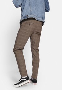Braune karierte Hose in Slim-Fit, mit einer Gesäßtasche und Knopfverschluss, kombiniert mit schwarz-weißen Sneakers. Eine Jeansjacke ist teilweise sichtbar.