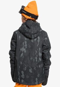 Svart jacka med camouflage-mönster och huva, strukturerad tyg och elastiska ärmslut. Kombination med en klar orange mössa och skidglasögon.