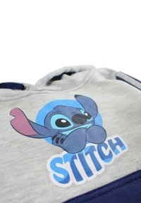 Sweat-shirt gris avec un motif Stitch bleu et le mot "STITCH" en blanc. Comprend une bande inférieure unie bleu marine et une texture douce.
