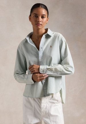 Donna con i capelli raccolti, che indossa una camicia verde chiaro con bottoni e pantaloni bianchi, mentre aggiusta il polsino su uno sfondo beige uniforme.