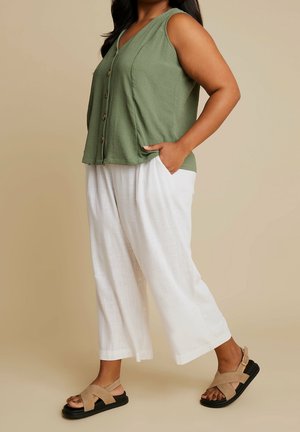 Woman wearing sleeveless olive green buttoned top, white wide-leg pants, and beige sandals with black soles against beige background.