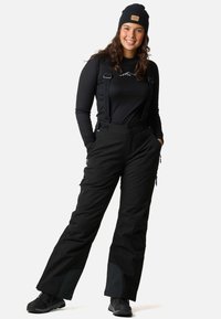 Zwarte skibroek met verstelbare banden, ritszakken en fleece details aan de boorden, gecombineerd met een zwart longsleeve en een beanie.