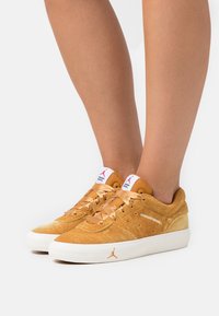 Bruna mockaskor med vita gummisulor. Har satängsnören och en präglad logotyp. Lågskärning med rund tå och perforerade detaljer.