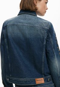 Denimjacka i mörkblått med en åtsittande design. Har broderade detaljer på ärmarna och en läderlogotyp på baksidan.