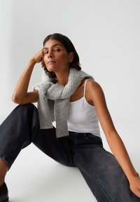 Woman seated with arm resting on knee, wearing white tank top, dark jeans, and a gray knit sweater draped over shoulders against plain background.