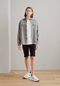 Camisa gris de botones con dos bolsillos en el pecho, llevada sobre una camiseta blanca, combinada con pantalones cortos negros y zapatillas blancas, en un fondo neutro.