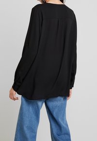 Black long-sleeve blouse with a relaxed fit and buttoned cuffs, featuring a smooth texture and a subtle back yoke design. Paired with light blue jeans.