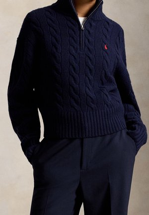 Marineblå kabelstrikket sweater med halvlukning og rødt logo på brystet, båret over en hvid skjorte, kombineret med marineblå bukser, hænderne i lommerne.