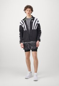 Veste de sport noire avec des rayures blanches, t-shirt gris, shorts noirs à motifs et baskets blanches. Comprend une fermeture éclair et des détails contrastants.