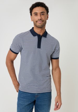 Uomo in piedi che indossa una polo a maniche corte a righe blu navy e bianco con colletto blu navy e jeans blu su sfondo uniforme.