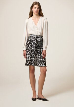 Blusa in tessuto crema con scollatura a V e maniche a sbuffo, abbinata a una gonna nera con motivo circolare bianco e vita elastica.