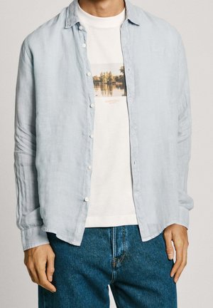 Mann trägt hellblaues Hemd mit Knöpfen über weißem T-Shirt mit Seemotiv, kombiniert mit blauen Jeans auf einfarbigem Hintergrund.